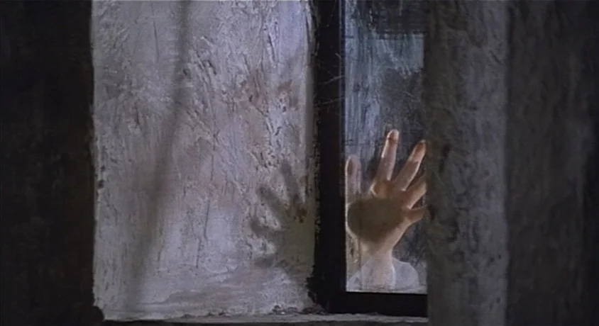 Kill, Baby Kill (1966) movie still: closeup — A dark, textured stone wall frames a single window pane at night. A pale hand presses against the…