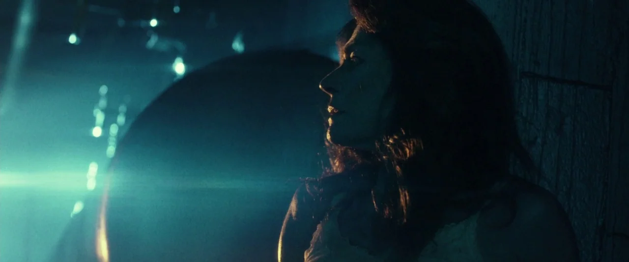 31 (2016) movie still: closeup — A dark space lit by high contrast teal light and lens flares. A woman is partially obscured by shadows and looks…