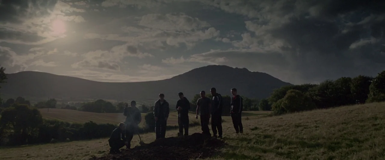 Kissing Candice (2017) movie still: wide — A group of seven men stands in a grassy field beneath a cloudy sky. One man kneels by a patch of freshly…