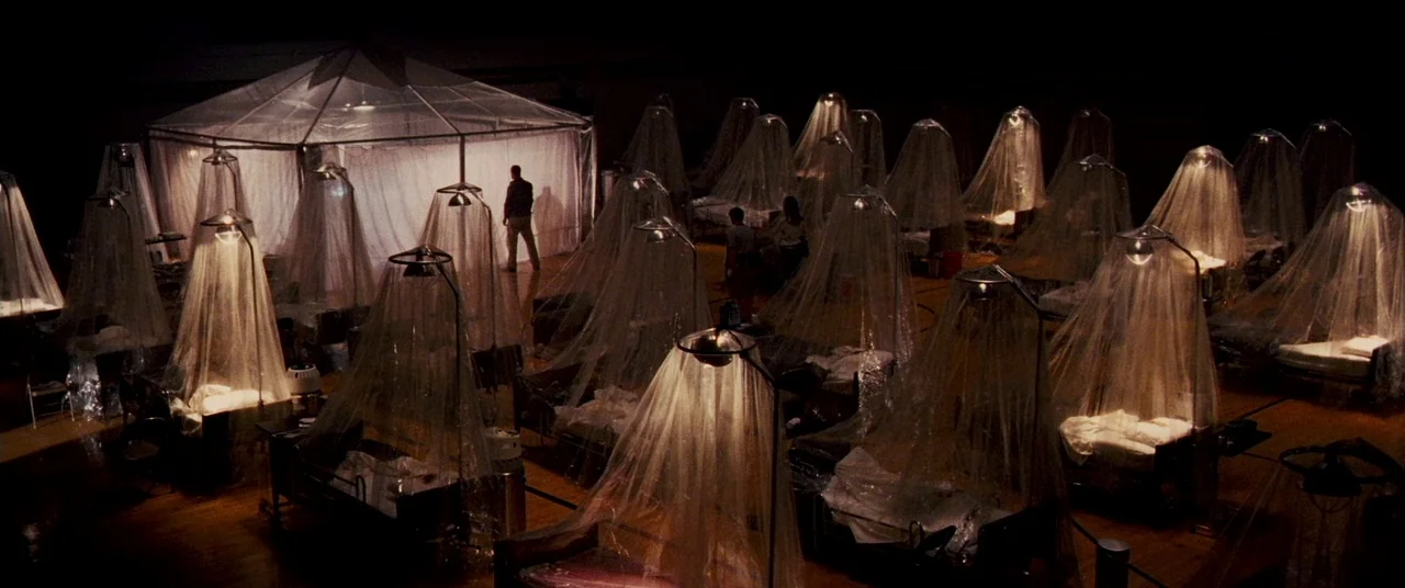 Carriers (2009) movie still: establishing — A dark gymnasium is filled with rows of cots draped in individual plastic tents. Small warm lamps…