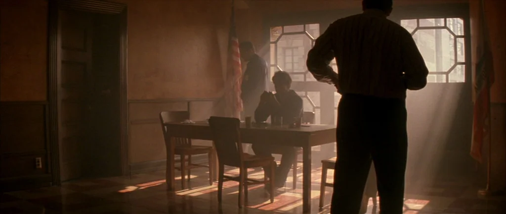 True Romance (1993) movie still: wide — A dimly lit room features a wooden table and several chairs under dusty sunlight. Three men stand or sit…