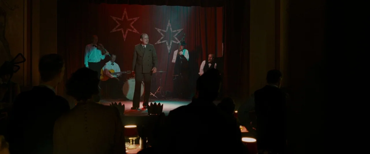 Phoenix (2014) movie still: wide — A dimly lit club features a performance stage framed by deep red velvet curtains. A middle-aged man in a suit…