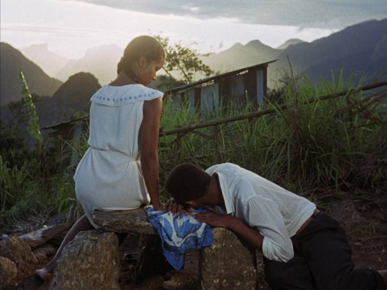 Black Orpheus (1959) movie still: fullbody — A woman in a white dress sits on a stone ledge on a mountainside. A man leans forward with his head…