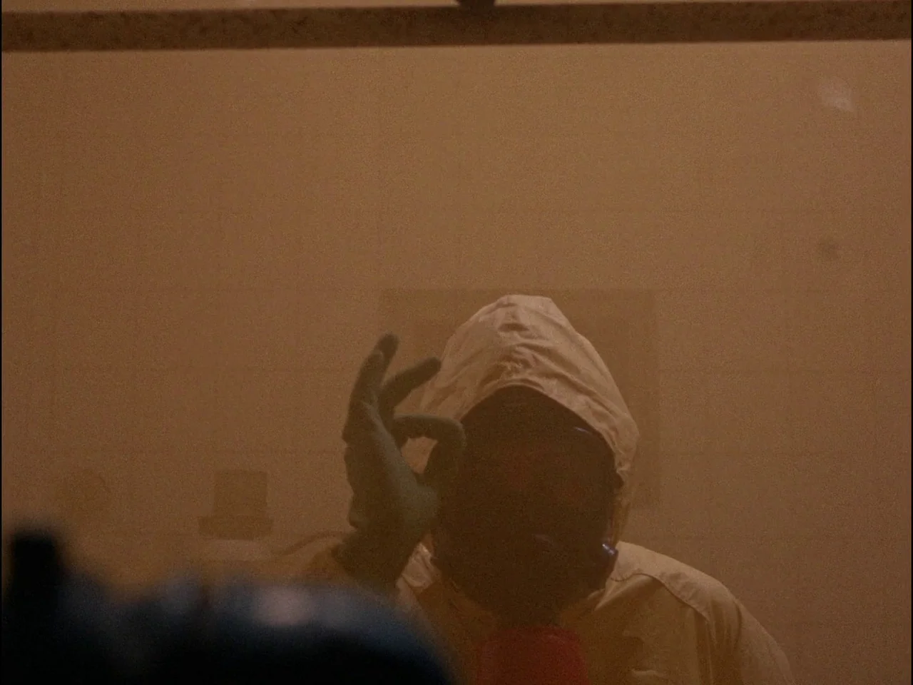 Dillinger is Dead (1969) movie still: closeup — A hazmat-suited figure stands in a haze-filled, dimly lit industrial lab environment. The subject…