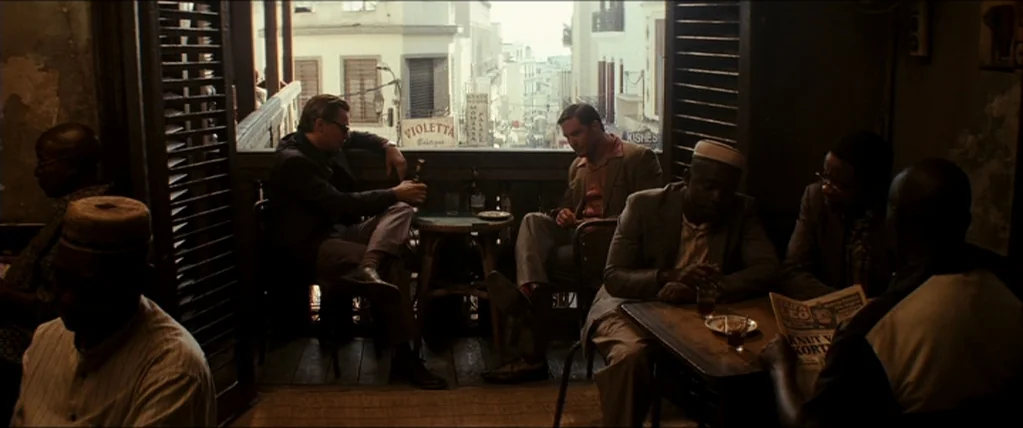 Inception (2010) movie still: wide — A dimly lit room with louvered wooden shutters opening onto a busy street scene. Several men sit at wooden…
