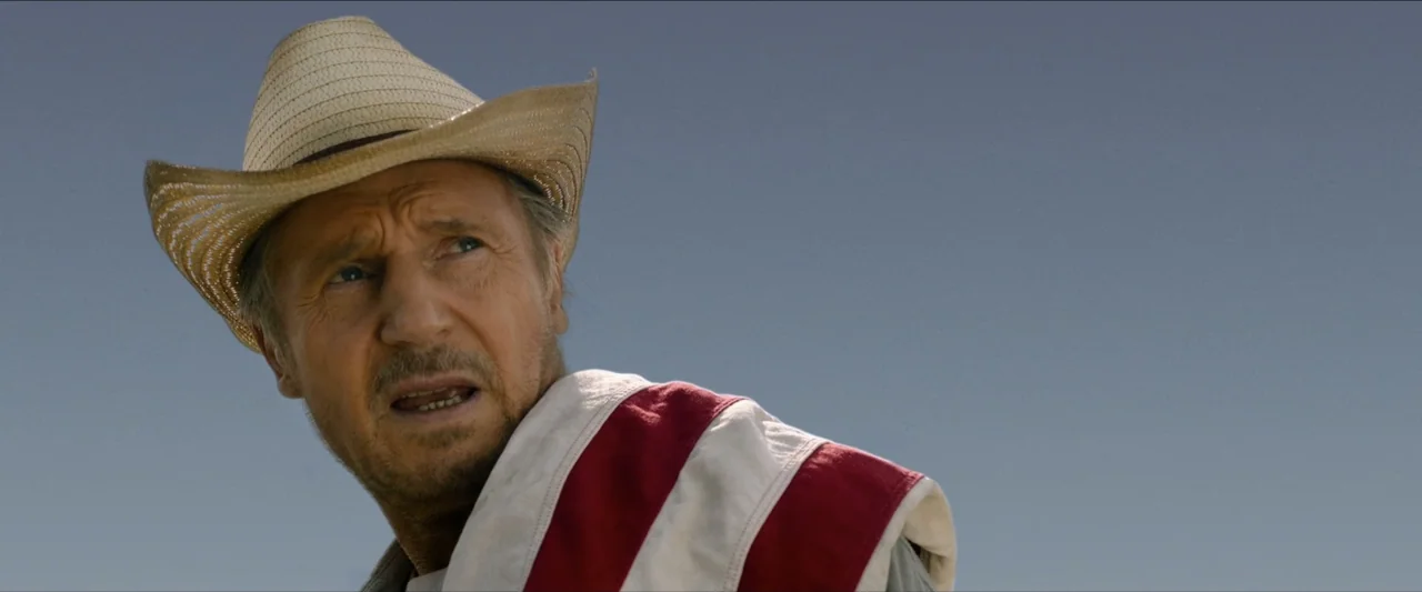 The Marksman (2021) movie still: closeup — A man stands in an arid desert environment under a clear blue sky. The man wears a wide-brimmed straw hat…