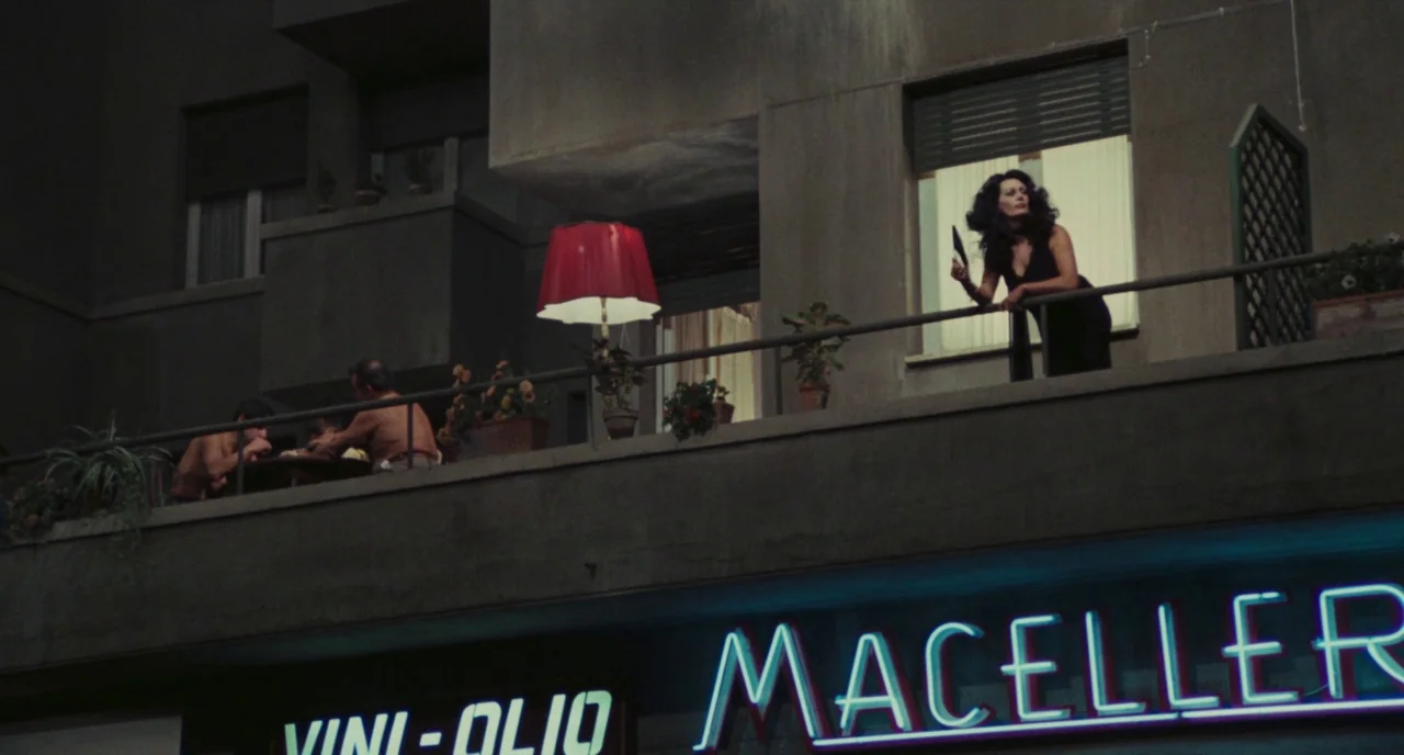 Roma (1972) movie still: wide — A concrete building exterior at night features a neon-lit shop sign below a balcony level. A woman stands by the…