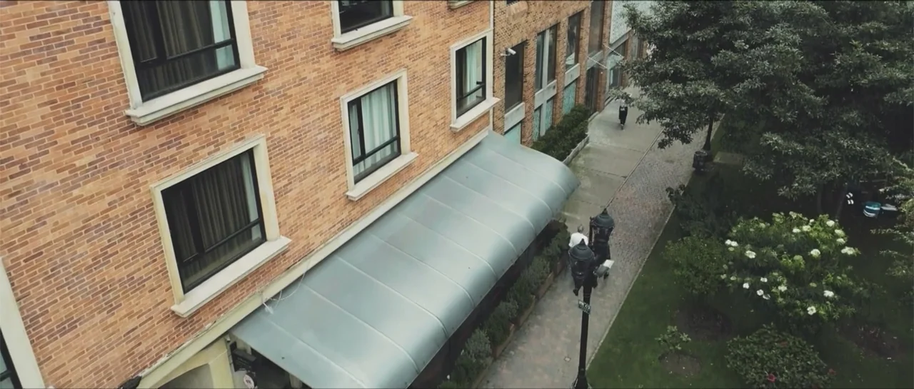 Loving is Losing (2019) movie still: establishing — A multi-story brick building features a metal awning over a narrow pedestrian walkway. Two…