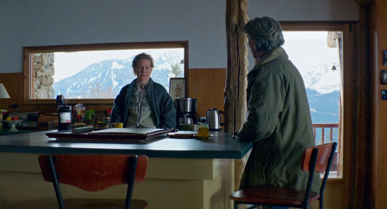 Anatomy of a Fall (2023) movie still: medium — A rustic kitchen featuring wooden paneling and a large window overlooking snowy mountains. A man and…