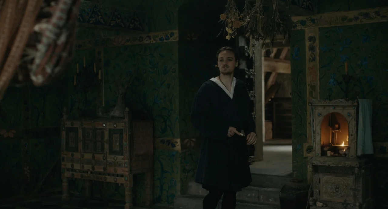 Catherine Called Birdy (2022) movie still: medium — A dimly lit room with walls painted in dark green floral patterns. A young man walks toward the…
