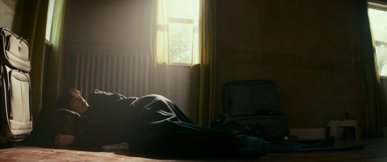Colossal (2016) movie still: medium — A dimly lit room features a hardwood floor and windows draped with yellow curtains. A person lies on the floor…