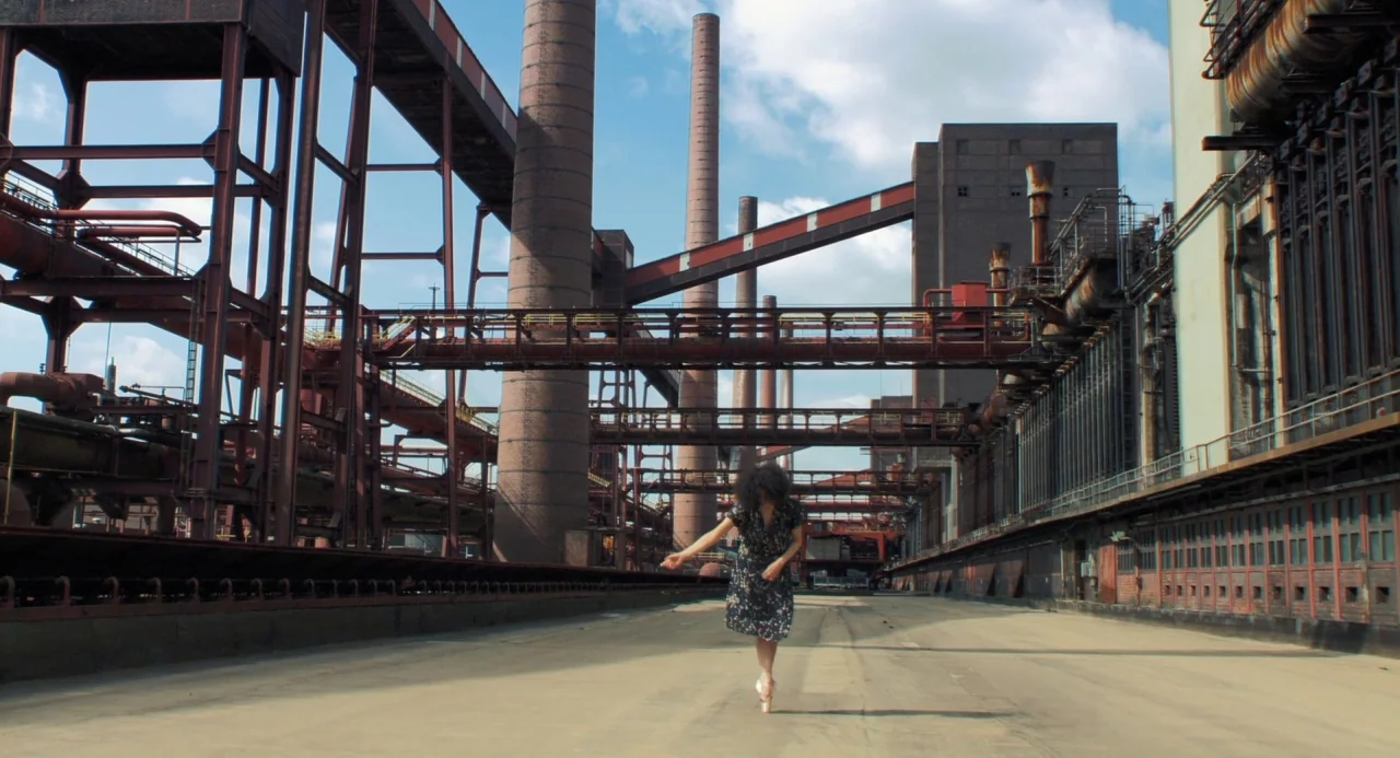 Pina (2011) movie still: wide — A vast, abandoned industrial site features towering red brick chimneys and steel structural scaffolding. A woman in…