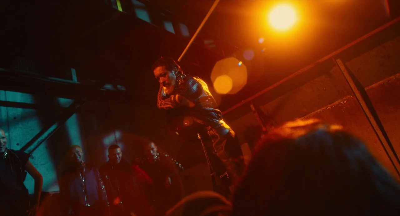 Diner (2019) movie still: medium — A dimly lit industrial nightclub space features heavy shadow and high contrast lighting. A bearded man in…