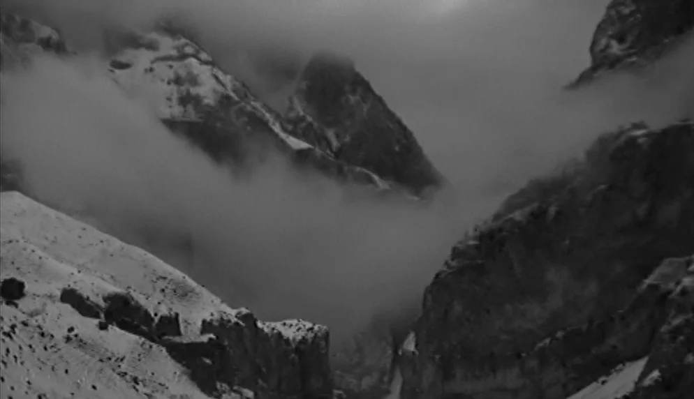 The Plea (1968) movie still: establishing — A desolate high-altitude mountain range features craggy rocks covered in patches of white snow. Thick,…