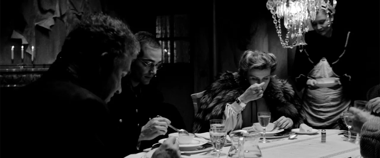 Europa (1991) movie still: medium — A dimly lit room features a dining table set for an evening meal. A small group of people sit together at the…