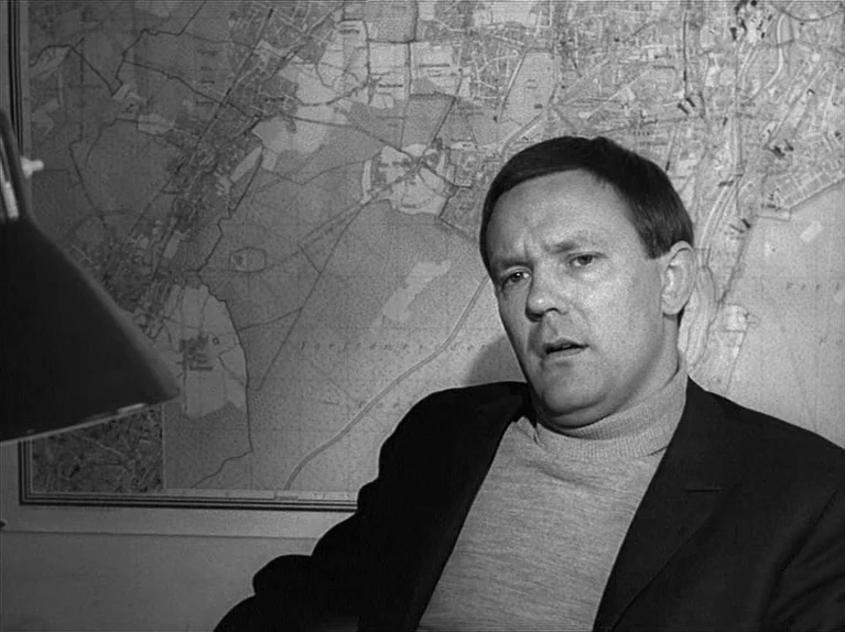 Gods Of The Plague (1970) movie still: medium — A man sits in front of a wall-mounted map in a dimly lit space. He looks toward the camera while…