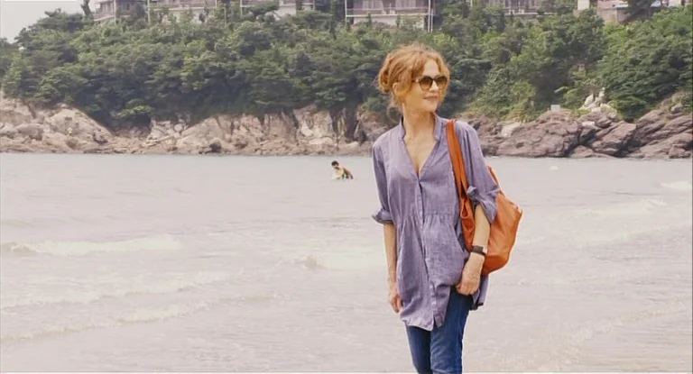 In Another Country (2012) movie still: fullbody — A sandy shoreline meets a rocky hillside topped with trees and houses. A woman in a blue tunic…