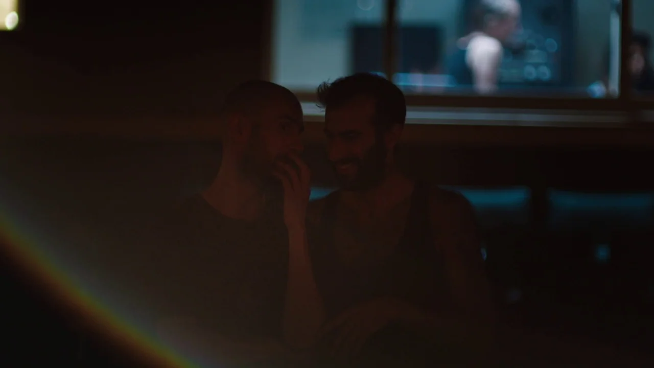 Madeline’s Madeline (2018) movie still: medium — A dark, dimly lit bar interior featuring two men sitting together near a window. One man whispers…