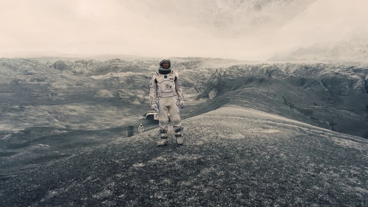 Interstellar (2014) movie still: fullbody — A vast, barren alien landscape with craggy grey terrain and distant jagged mountains. An astronaut…