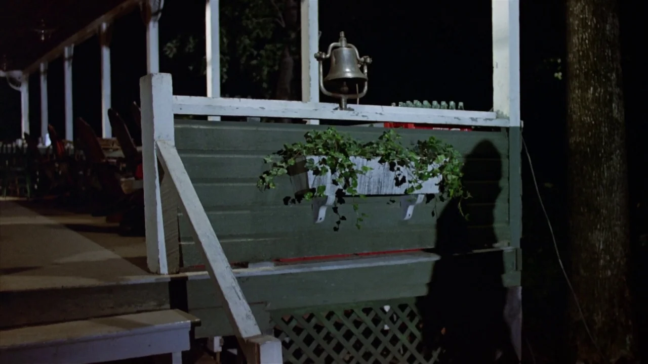 Friday the 13th Part 2 (1981) movie still: wide — A wooden porch at night features a green-painted railing and a white lattice base. An iron bell…