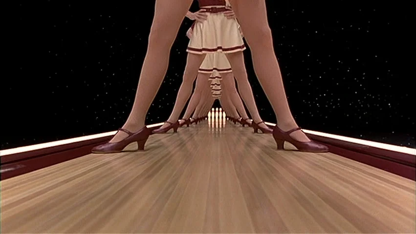 The Big Lebowski (1998) movie still: closeup — A polished wooden bowling lane stretches toward a set of white bowling pins. Endless rows of legs…
