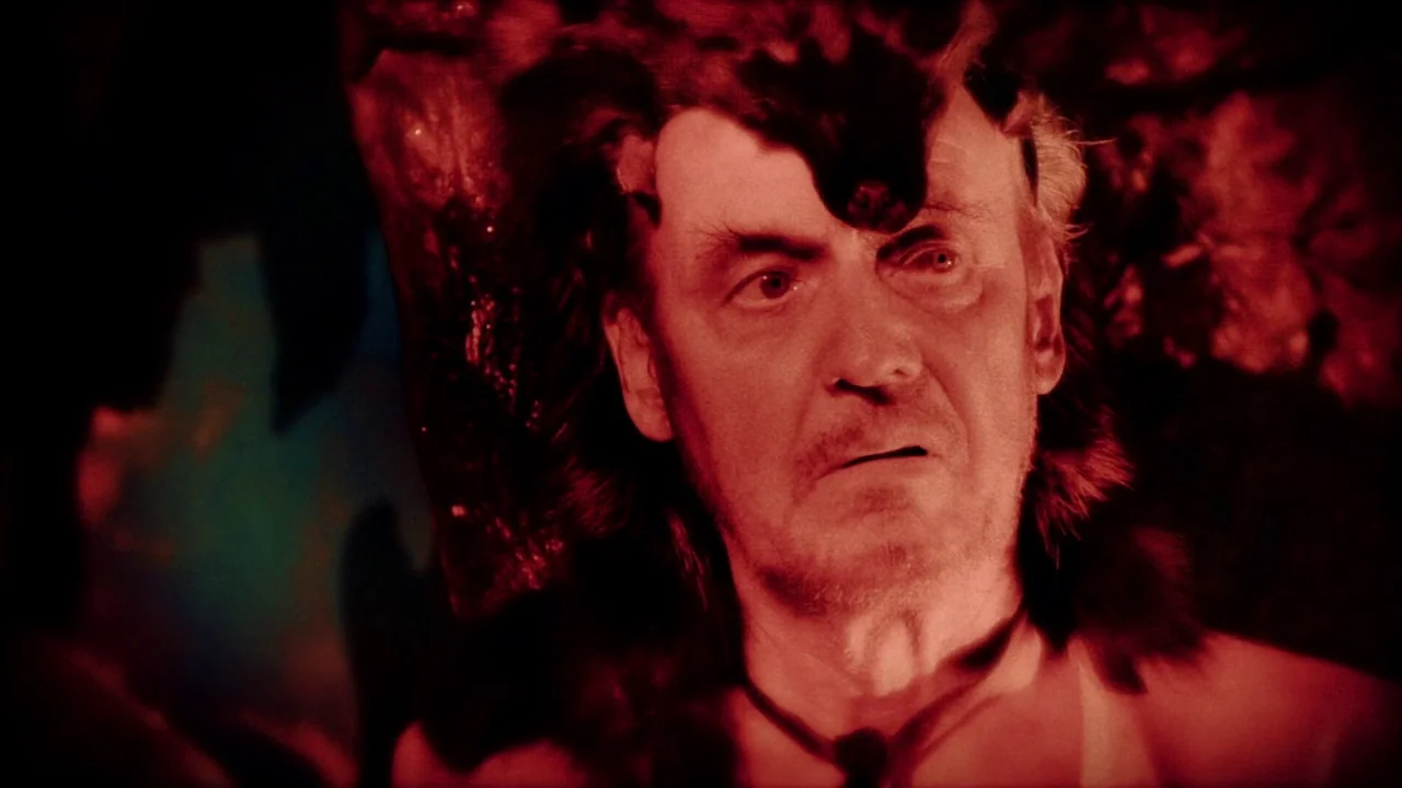 The Forbidden Room (2015) movie still: closeup — A dark cave interior is bathed in an intense and saturated red light. An older man with grey hair…