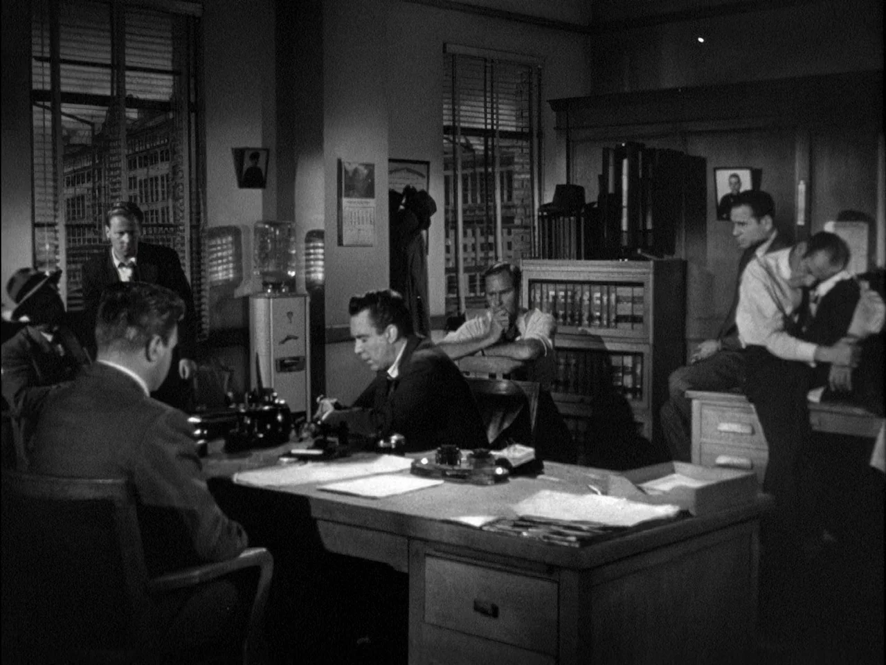 D.O.A. (1949) movie still: wide — A crowded professional office features wooden desks, bookshelves, and large windows with blinds. Multiple men in…