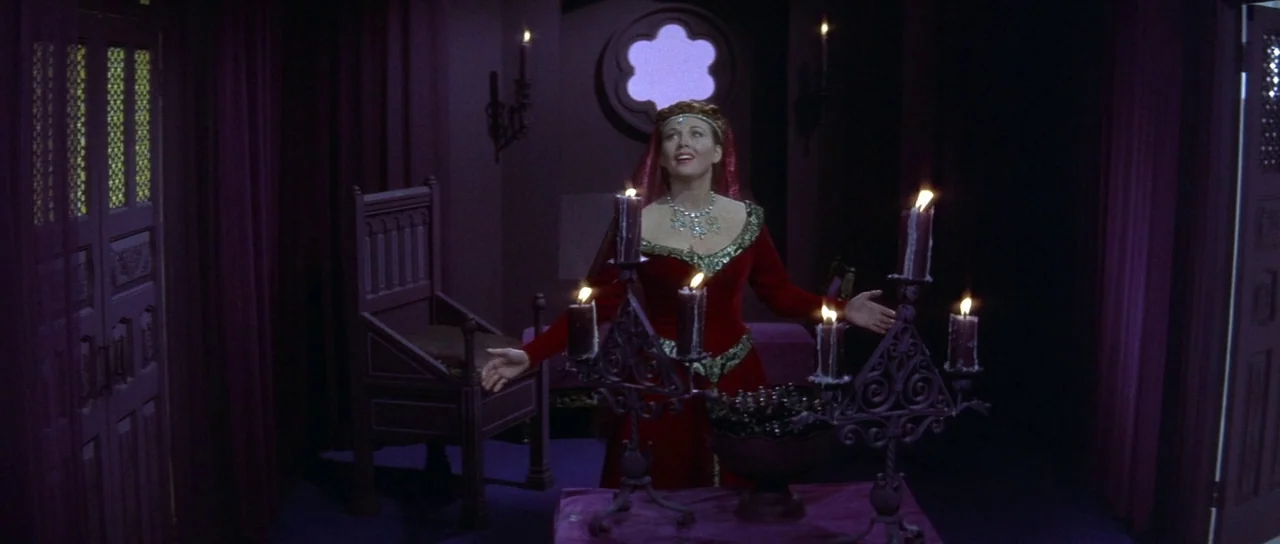 The Masque of the Red Death (1964) movie still: medium — A dimly lit room with purple velvet curtains and ornate wooden furnishings. A woman stands…