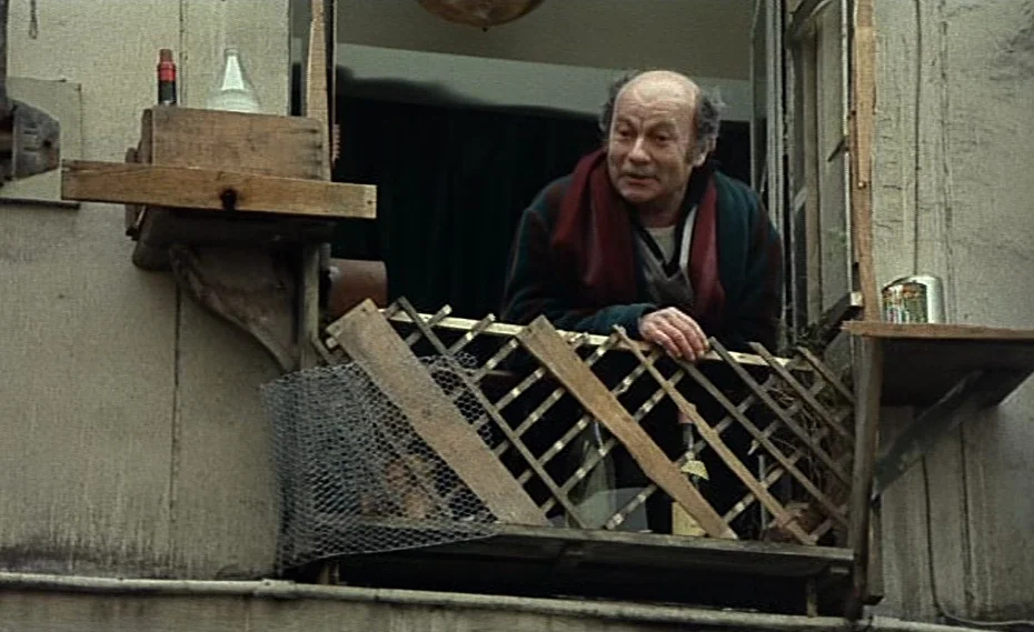 Bed & Board (1970) movie still: medium — An aging man stands on a makeshift wooden balcony attached to an old building facade. The man leans on a…