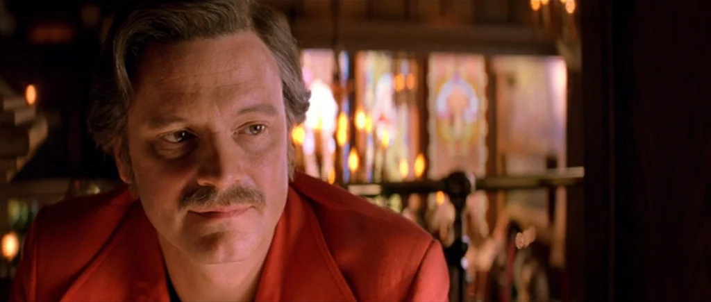 Where The Truth Lies (2005) movie still: closeup — A dark indoor space features blurred decorative stained glass in the background. A man in a red…