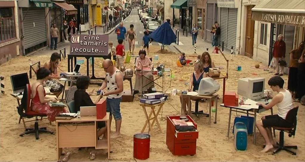 The Beaches of Agnès (2008) movie still: wide — An urban French street is covered in a thick layer of sand. Several individuals sit at office desks…