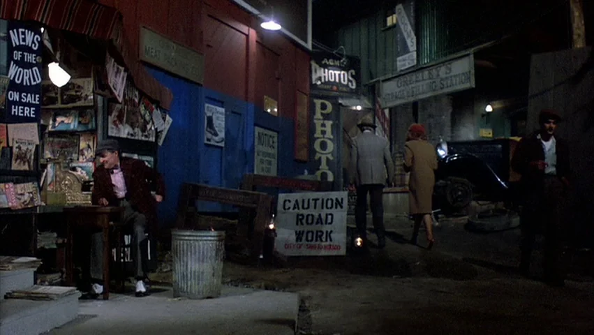 Hammett (1982) movie still: wide — A nighttime urban street corner features a newsstand kiosk and varied advertising signage. Two individuals walk…