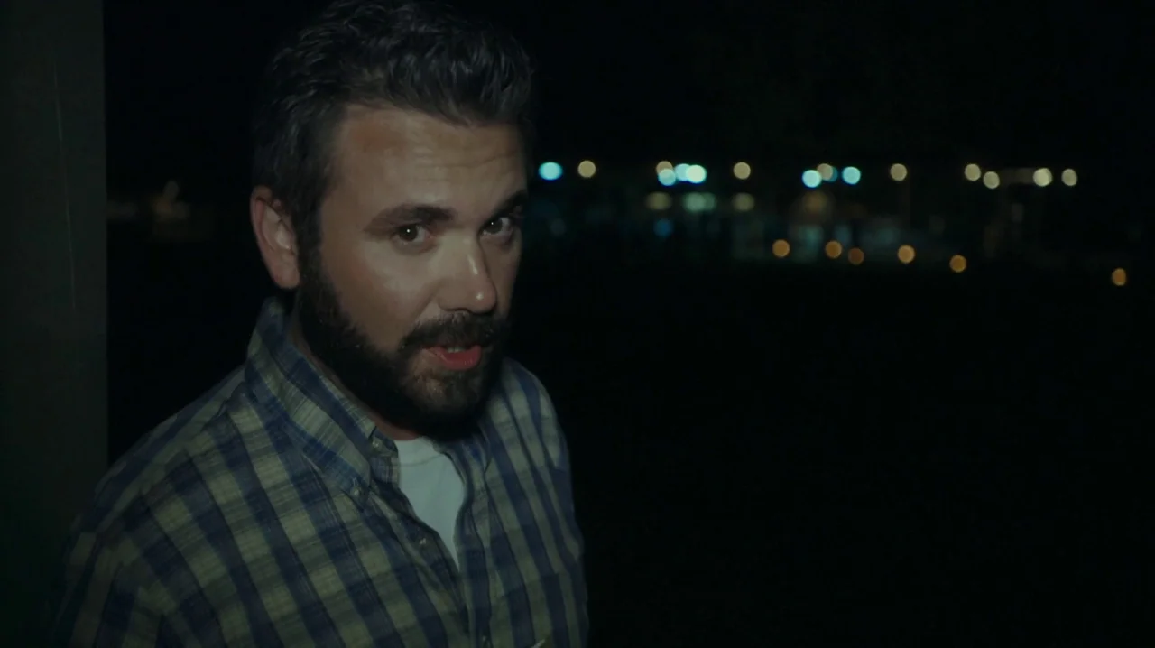 The Sacrament (2013) movie still: medium — An outdoor location at night features a dark foreground with blurred distant lights. A man with a dark…