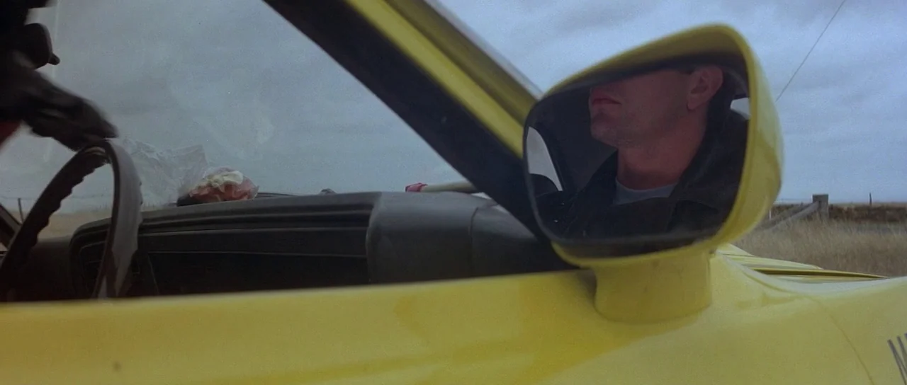 Mad Max (1979) movie still: closeup — A man's face is visible in the side mirror of a bright yellow car. The vehicle is parked or driving through a…