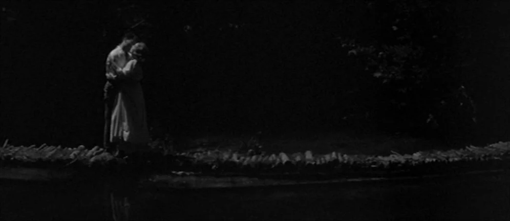 Les Amants (1958) movie still: wide — A rustic log footbridge spans a dark pool of water at night. A young couple in light clothing stands close…