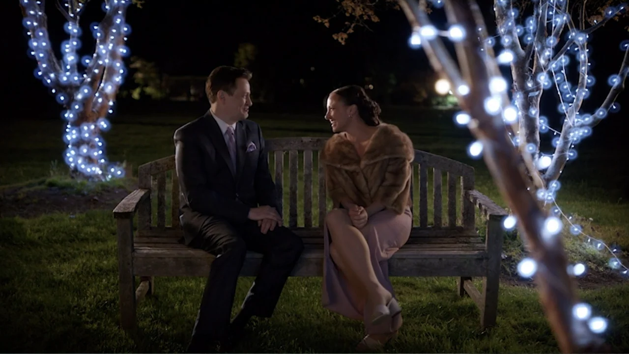 Ask Me to Dance (2022) movie still: medium — A wooden bench sits in a dark grassy area at night illuminated by string lights. A man in a dark suit…