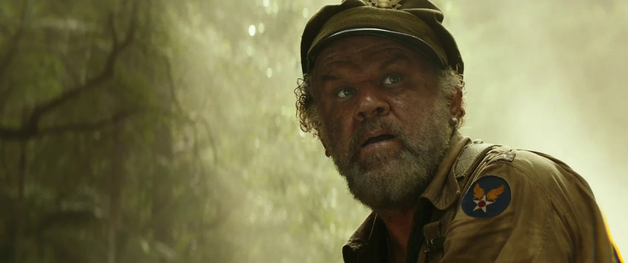 Kong: Skull Island (2017) movie still: closeup — A dense jungle environment with soft sunlight streaming through blurry trees and foliage. An older…