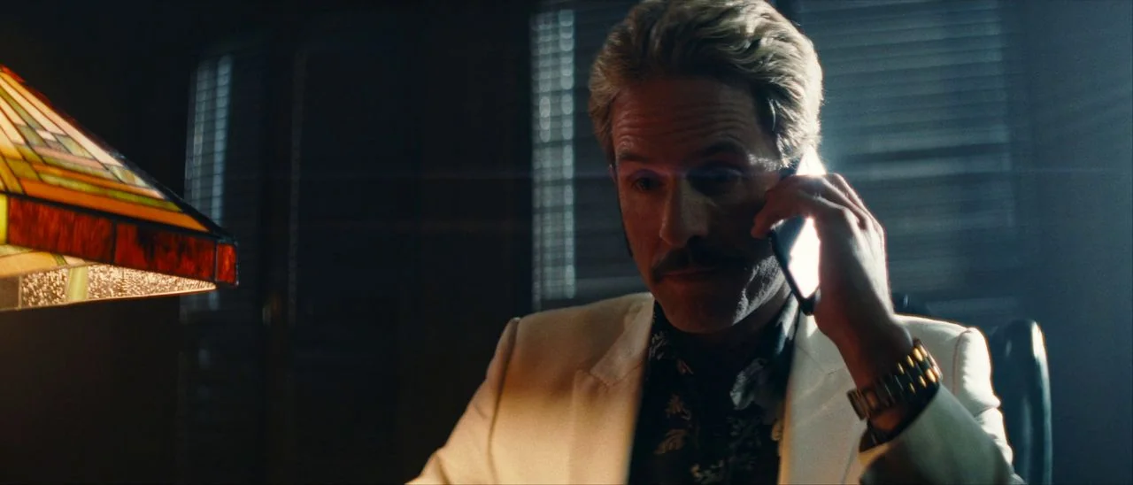 Archenemy (2020) movie still: medium — A man sits at a desk at night with window blinds visible behind him. He holds a smartphone to his ear while…