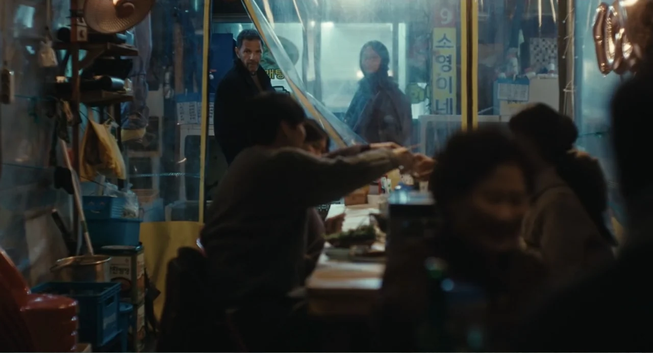Winter in Sokcho (2025) movie still: medium — A dimly lit street food stall is enclosed by translucent yellow and clear plastic sheeting. A man in a…