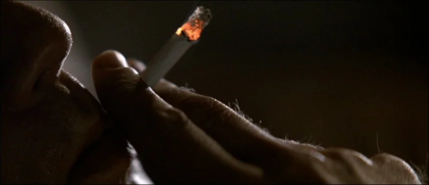 Road To Perdition (2002) movie still: closeup — A dark, enclosed space with minimal illumination. A hand holds a lit cigarette up to a person's mouth.