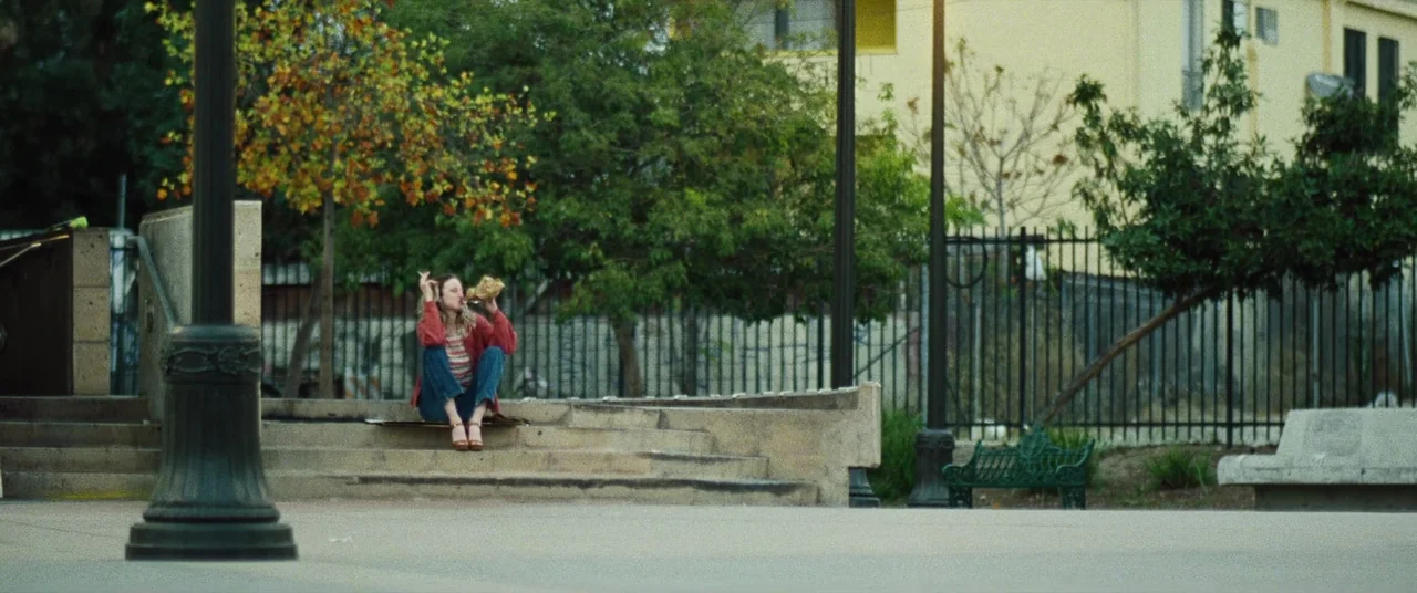 To Leslie (2022) movie still: wide — A public park area features concrete steps leading to a paved space with trees. A young woman sits on the steps…