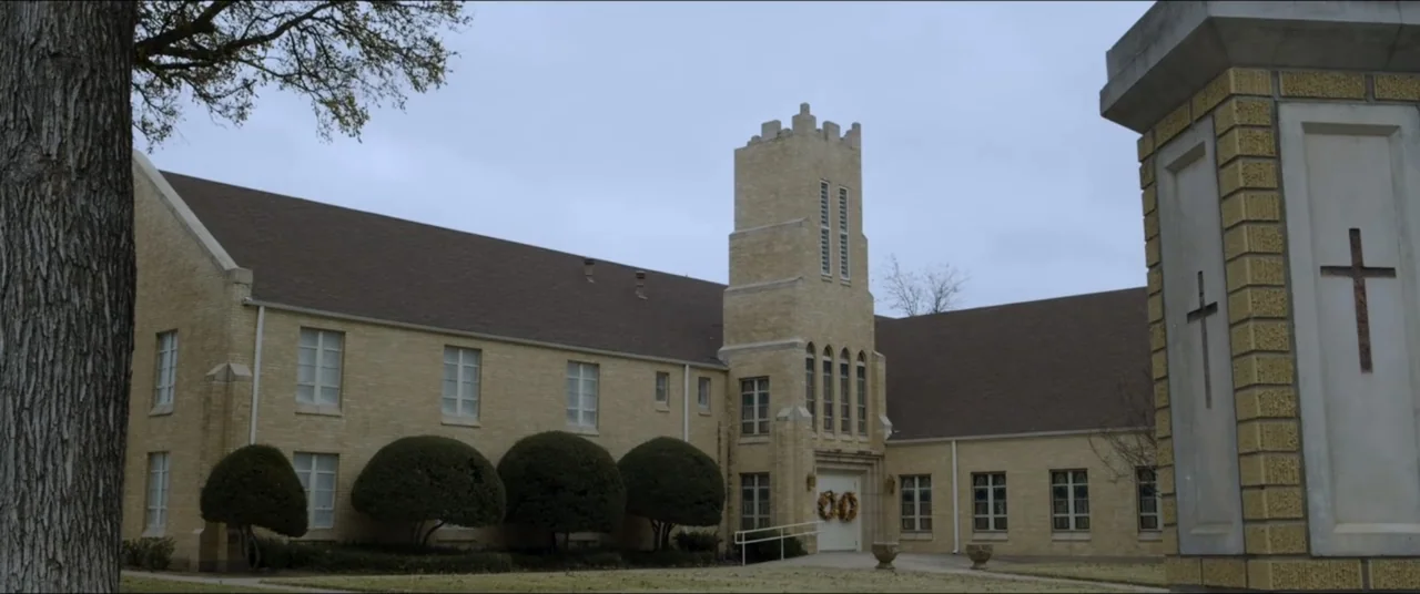 Amityville Cult (2021) movie still: establishing — A light-colored brick church building features a central square tower and a peaked roof. The…