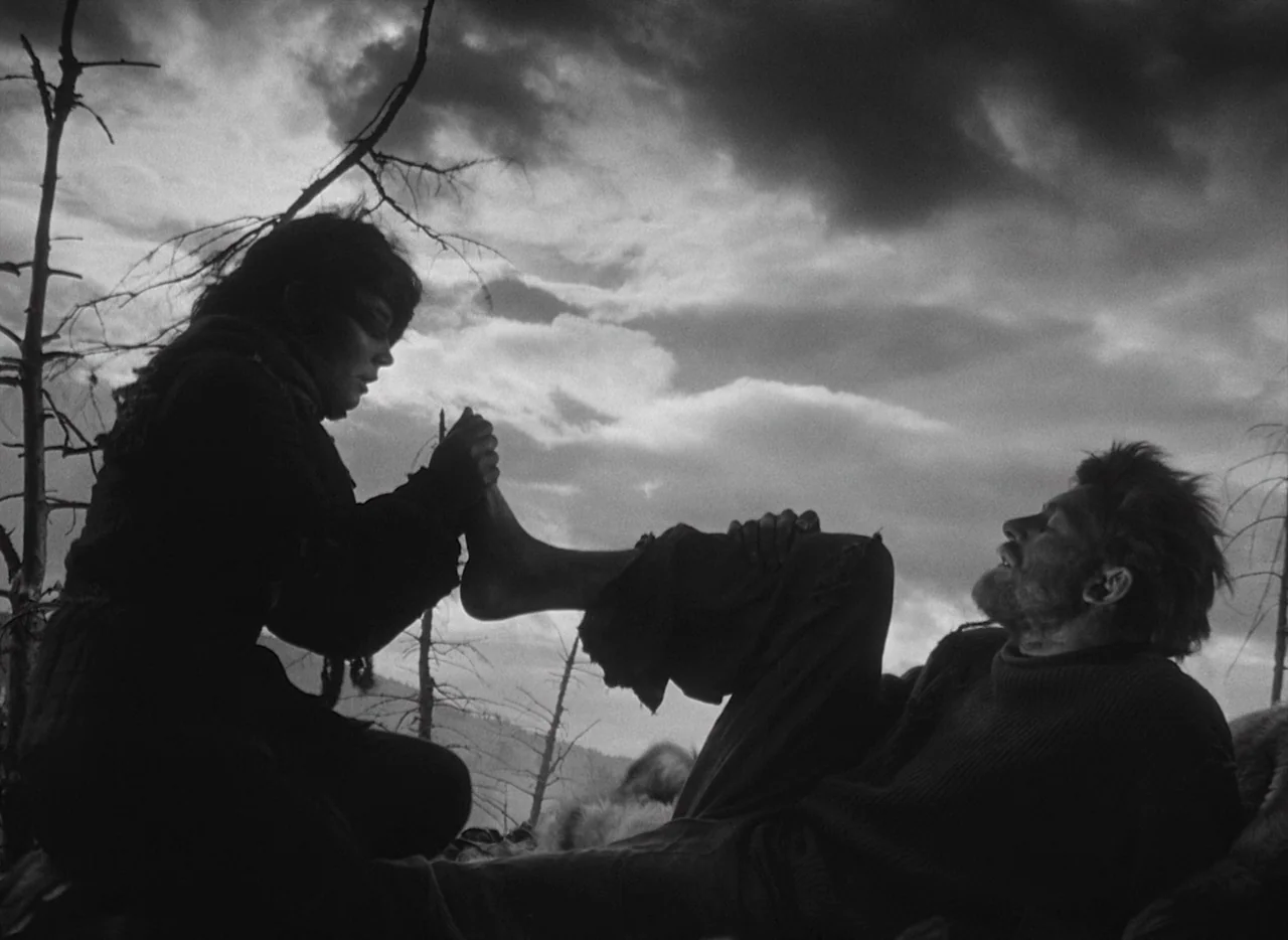 Letter Never Sent (1960) movie still: medium — A bleak, desolate hillside with skeletal trees under a heavy, overcast, dramatic sky. A woman sits on…
