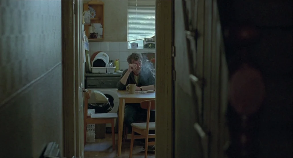 Looking for Eric (2009) movie still: wide — A small, cluttered kitchen seen through a narrow doorway from an adjacent hallway. A man sits at a small…