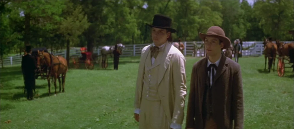 Ride With The Devil movie still: fullbody — A sunny clearing surrounded by dense trees with a white wooden fence in back. Two men in 19th-century…