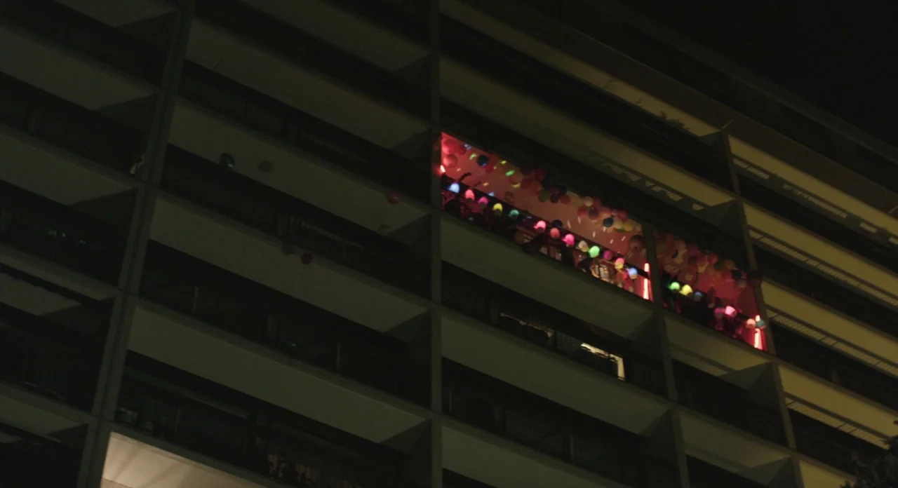 Holy Motors (2012) movie still: wide — A dark apartment building facade features one illuminated balcony filled with colorful party decorations.…