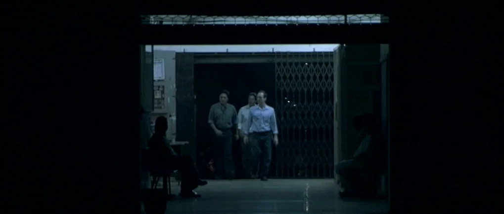 A Mighty Heart (2007) movie still: wide — A dimly lit indoor corridor with an open metallic gate at the far end. Three men walk side by side towards…