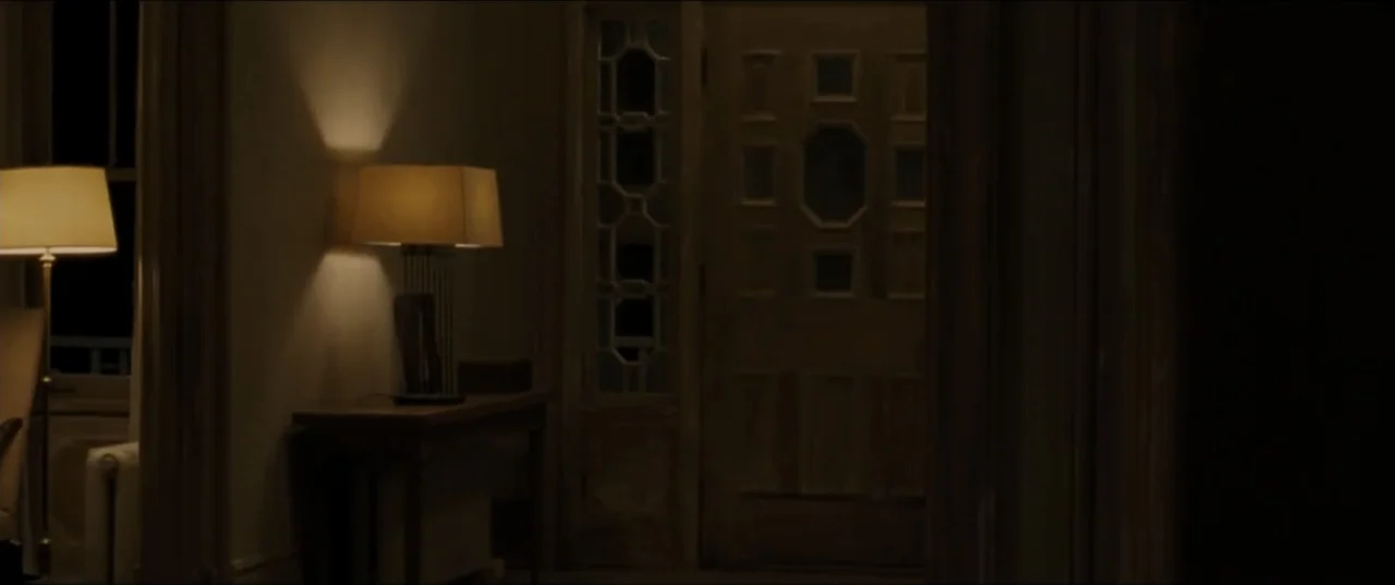 mother! (2017) movie still: wide — A dimly lit residential hallway at night features a wooden door and table lamps. An empty corridor remains silent…
