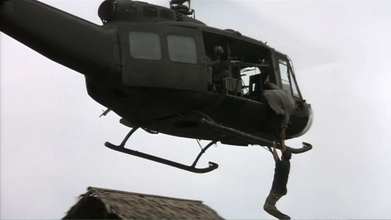 Behind Enemy Lines (2001) movie still: wide — A military-style helicopter hovers mid-air above a thatched roof structure in an open landscape. One…