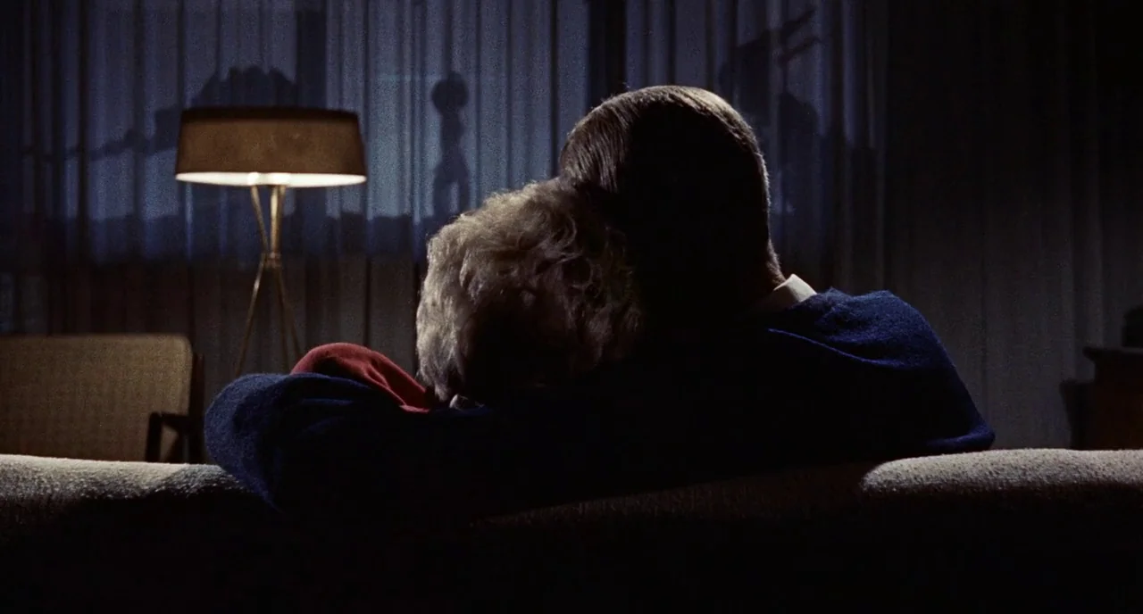 Bell Book and Candle (1958) movie still: medium — A dimly lit room with a sofa facing a window covered by curtains. A couple sits together in the…